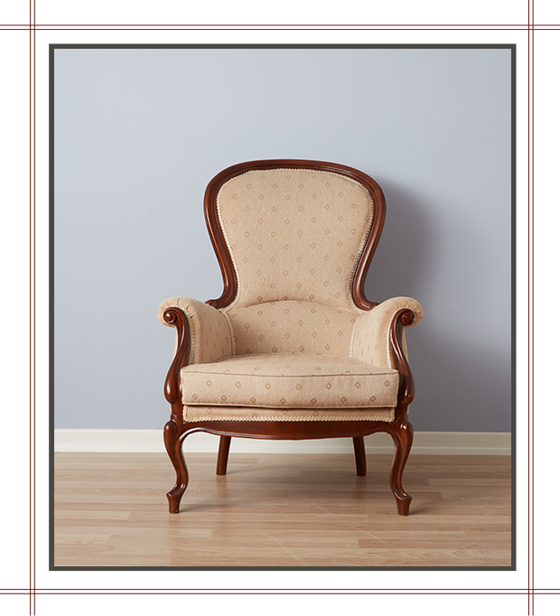 An ornate beige upholstered armchair with wooden trim placed on a light wood floor against a light gray wall.